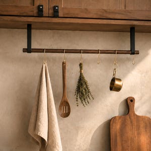 Puede incluir: Un estante de almacenamiento de cocina con una barra de madera oscura y soportes de metal negro, montado debajo de un armario. Ganchos dorados sostienen una toalla beige, una cuchara de madera, hierbas y una pequeña taza medidora de latón. También se ve una tabla de cortar de madera.
