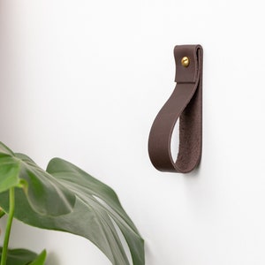 Op de afbeelding: Een bruine leren haak met een messing schroef, gemonteerd op een witte muur. De haak is lusvormig en is ontworpen om items aan op te hangen.