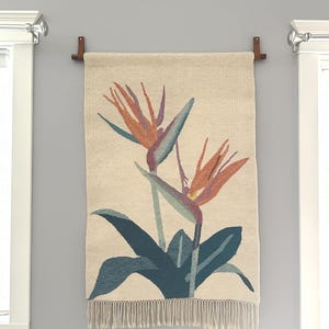 May include: Woven wall hanging featuring a bird of paradise flower design. The artwork showcases orange and blue flowers with green leaves against a cream-colored background. The bottom edge has a fringe.