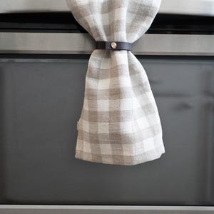 Modern leather towel holder, also called a towel hugger, securing a neutral plaid kitchen towel on an oven handle with brass hardware.