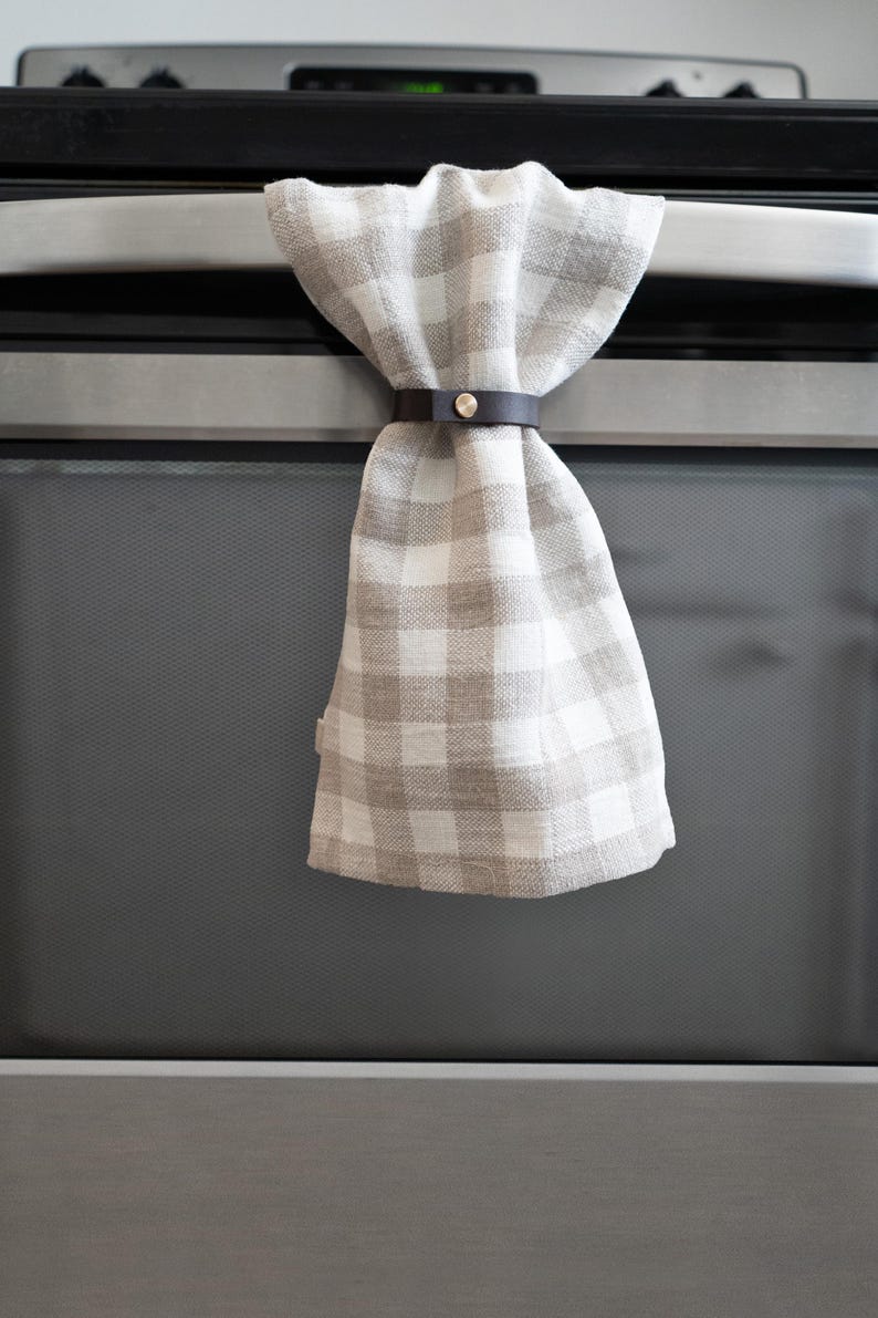 Modern leather towel holder, also called a towel hugger, securing a neutral plaid kitchen towel on an oven handle with brass hardware.