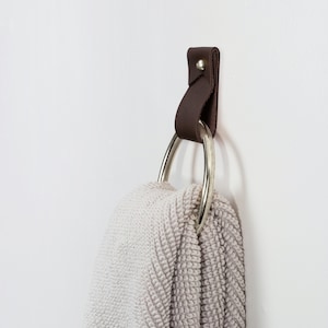 May include: A brown leather towel ring with a silver metal ring. The towel ring is mounted on a white wall and has a towel hanging from it.