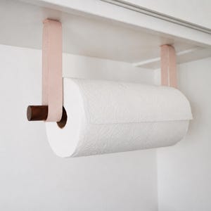 以下が含まれることがあります： 白いペーパータオルのロールが付いたペーパータオルホルダー。ホルダーはキャビネットに取り付けられており、薄いピンクのストラップと濃い茶色の木製ダボが付いています。ホルダーはキャビネットの下に取り付けるように設計されています。