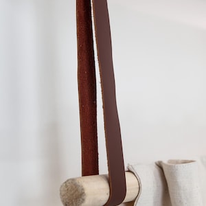 May include: A brown leather strap is attached to a light-colored wooden dowel, designed for hanging items. The strap is suspended from above, and a light-colored fabric is draped over the dowel. The design is simple and minimalist.