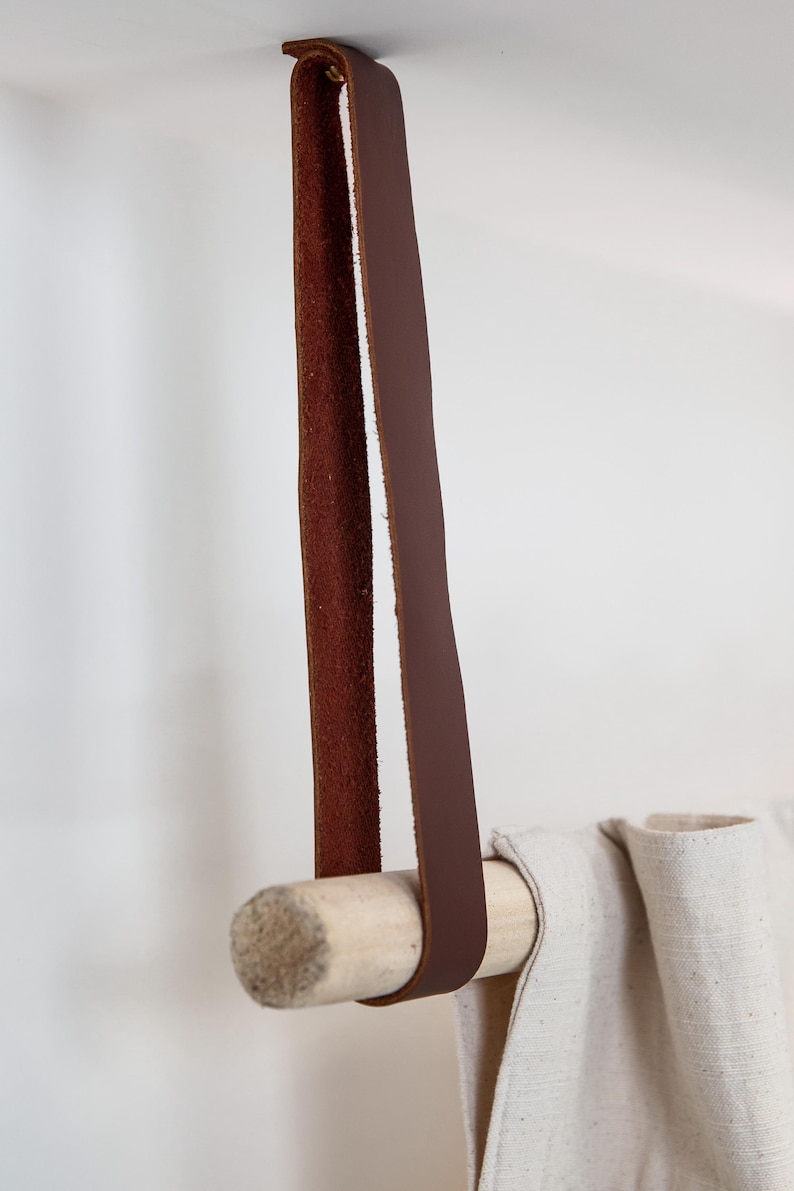 May include: A brown leather strap is attached to a light-colored wooden dowel, designed for hanging items. The strap is suspended from above, and a light-colored fabric is draped over the dowel. The design is simple and minimalist.