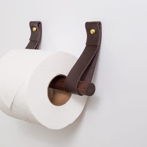 May include: A modern toilet paper holder with a roll of white paper. The holder features dark brown leather straps attached to the wall with brass rivets. A wooden dowel holds the paper roll.