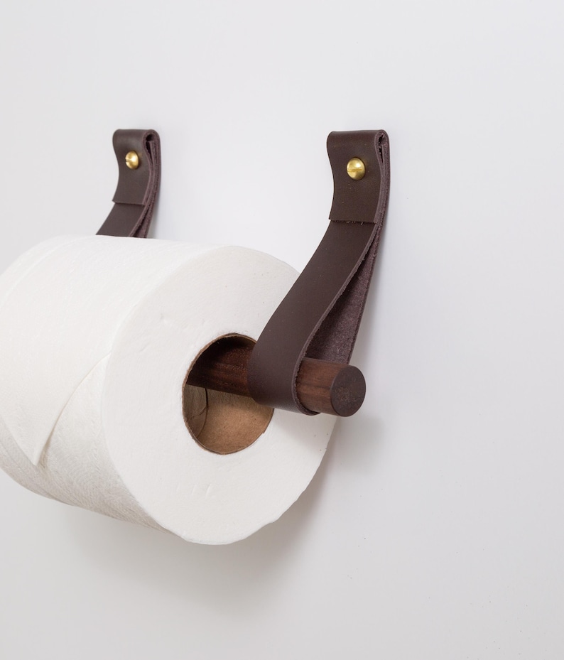 May include: A modern toilet paper holder with a roll of white paper. The holder features dark brown leather straps attached to the wall with brass rivets. A wooden dowel holds the paper roll.