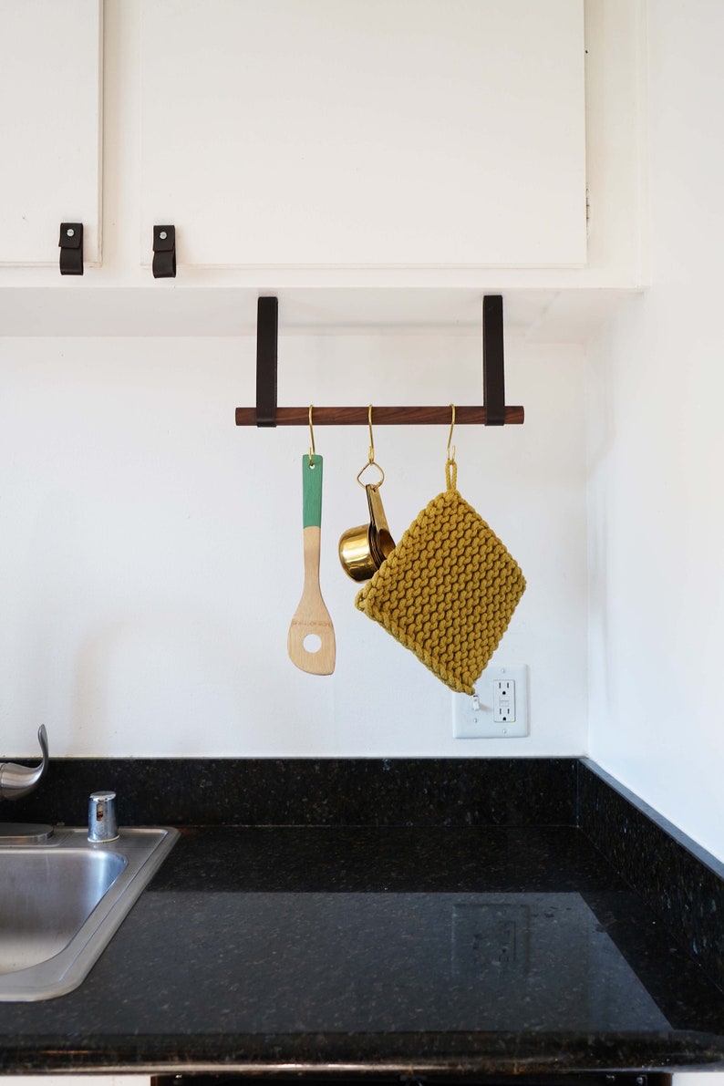 May include: A wooden kitchen utensil rack with three hooks. A green wooden spatula, a gold measuring cup, and a yellow knitted pot holder are hanging from the hooks.