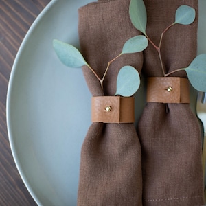 May include: Two brown linen napkins with leather napkin rings and sprigs of greenery on a light blue plate.