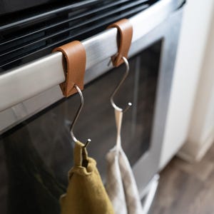 May include: Two brown leather oven rack hooks with stainless steel S-hooks, holding two kitchen towels. The hooks are attached to the oven handle. One towel is olive green, the other is off-white.