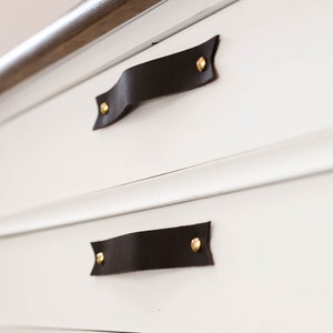 Angled close-up of Keyaiira Black genuine leather cabinet and drawer handles with V-shaped pointed ends and polished brass mounting hardware installed on white furniture, highlighting the deep Black leather, raised grip, natural texture and handcrafted construction