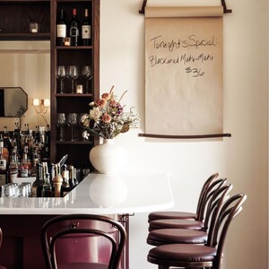 May include: A bar scene featuring a wooden wine rack with bottles and glasses, a white countertop, and maroon bar stools. A paper scroll with handwritten text, "Tonight's Special: Blackened Mahi-Mahi $36," hangs on the wall. A vase of flowers sits on the counter.