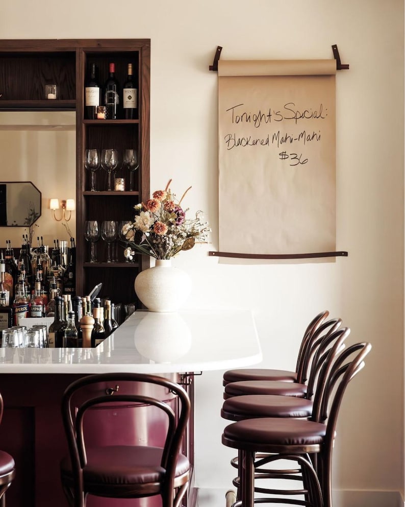 May include: A bar scene featuring a wooden wine rack with bottles and glasses, a white countertop, and maroon bar stools. A paper scroll with handwritten text, "Tonight's Special: Blackened Mahi-Mahi $36," hangs on the wall. A vase of flowers sits on the counter.
