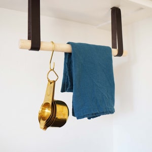May include: A wooden rod with two brown leather straps hanging from a white cabinet. A blue dish towel is hanging on the rod. A set of gold measuring cups is hanging from a hook on the rod.