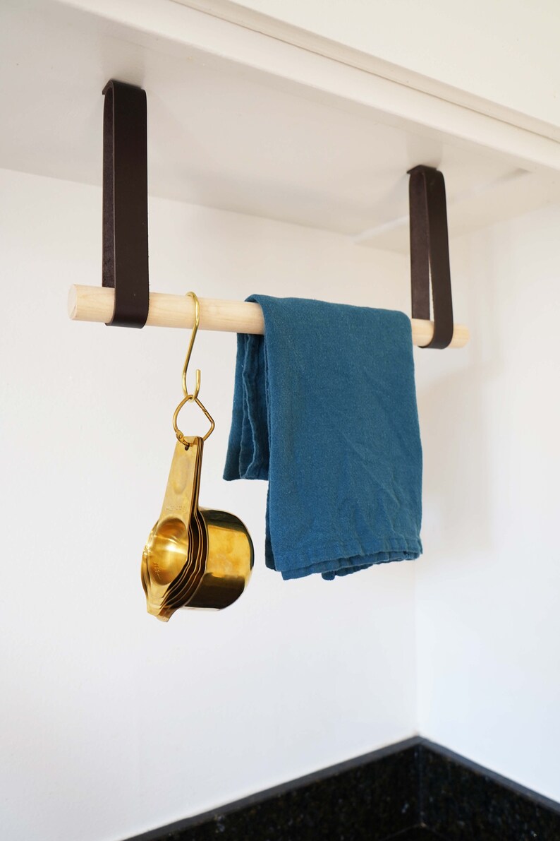 May include: A wooden rod with two brown leather straps hanging from a white cabinet. A blue dish towel is hanging on the rod. A set of gold measuring cups is hanging from a hook on the rod.