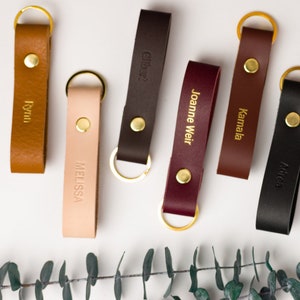 May include: A selection of personalised leather keyrings in shades of tan, blush, brown, burgundy, and black. Each keyring has a gold-coloured metal ring and a brass stud. Names are embossed in gold lettering on each strap.