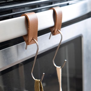 May include: Two brown leather and stainless steel S-hooks hanging from a stainless steel oven handle. The hooks are designed to hold kitchen towels or other items. The hooks are attached to the oven handle with a leather loop.