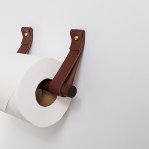 May include: A brown leather and wood toilet paper holder with a roll of white toilet paper. The holder features two leather straps attached to the wall with brass rivets. The wooden dowel holds the toilet paper roll.