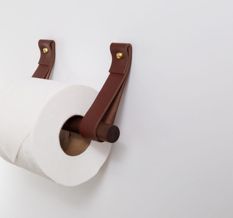 May include: A brown leather and wood toilet paper holder with a roll of white toilet paper. The holder features two leather straps attached to the wall with brass rivets. The wooden dowel holds the toilet paper roll.