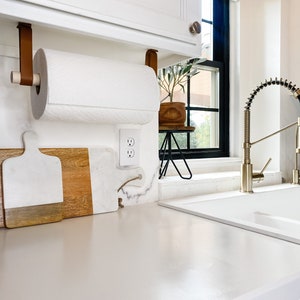 May include: A white kitchen countertop with a brown leather paper towel holder attached to a white cabinet above. The paper towel holder is holding a white paper towel roll. There are two white and brown cutting boards on the countertop. A white electrical outlet is visible on the wall. A small potted plant is on a black metal stand next to the outlet. A white kitchen sink is in the background.