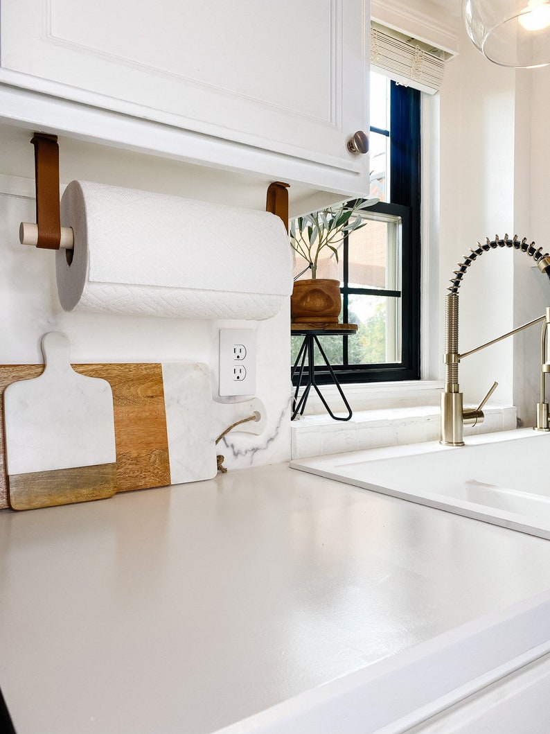 May include: A white kitchen countertop with a brown leather paper towel holder attached to a white cabinet above. The paper towel holder is holding a white paper towel roll. There are two white and brown cutting boards on the countertop. A white electrical outlet is visible on the wall. A small potted plant is on a black metal stand next to the outlet. A white kitchen sink is in the background.