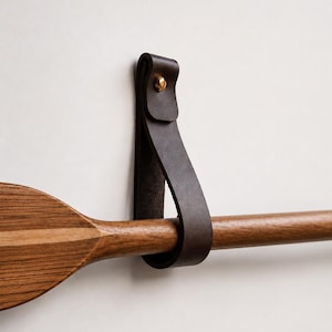 Peut inclure: Une pagaie en bois avec une sangle en cuir marron foncé et un rivet en laiton. La pagaie est faite de bois clair et foncé. La sangle en cuir est fixée au mur, maintenant la pagaie en place.