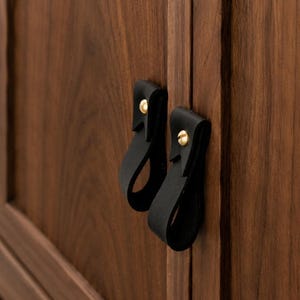 May include: Close-up of a wooden cabinet with two black leather pull handles. The handles are attached with gold-colored screws. The cabinet is made of dark brown wood with visible grain.