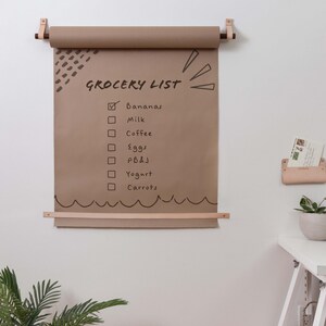 Keyaiira wall mounted kraft paper holder with vegetable tanned leather straps and wood dowels, displaying a reusable grocery list, family command center, kitchen organization board, homeschool planner, and modern farmhouse wall decor.
