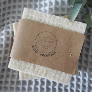 Op de afbeelding: Twee witte wollen sponzen met een bruin papieren etiket waarop "WOOL SPONGES" staat en een lijntekening van een schapenkop in een cirkel.
