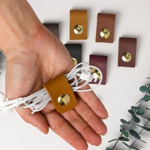 May include: A collection of leather cable organizers in various colors, including brown, black, and burgundy, each with a gold-colored snap closure. One organizer holds white earbuds. The organizers are displayed on a white surface with green foliage.