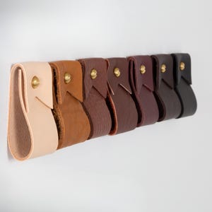 May include: A row of seven leather drawer pulls in varying shades of brown, from light tan to black. Each pull is attached with a brass rivet. The pulls are mounted on a white surface, showcasing the range of colors and textures.