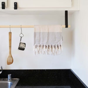 May include: A wooden kitchen towel rack with black metal brackets mounted under a white cabinet. A gray and white kitchen towel hangs on the rack. A wooden spoon and a metal strainer hang from hooks on the rack.