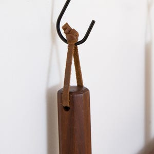 May include: A dark brown wooden handle with a leather strap is suspended from a black metal hook. The handle has a geometric shape and a hole near the top. The leather strap is knotted at the top.