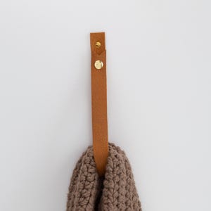 May include: A brown leather strap with gold-colored rivets is mounted on a white wall, holding a brown knitted item. The strap is rectangular and appears to be made of genuine leather. The knitted item has a chunky texture.