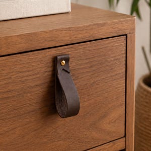 May include: Close-up of a wooden nightstand with a dark brown leather pull. The nightstand is made of light brown wood and has two drawers. A white ceramic vase and a book are on top.