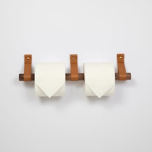 May include: A double toilet paper holder with a minimalist design. The holder features two rolls of white toilet paper suspended by a dark brown wooden rod and secured with tan leather straps and brass rivets. The fixture is mounted on a white wall.