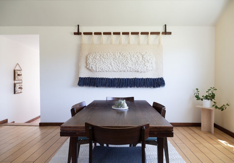 May include: A white and blue woven wall hanging with a fringe border hangs above a dark wood dining table with four chairs.