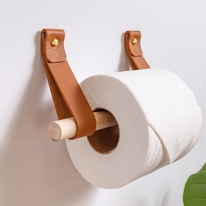 May include: A wooden toilet paper holder with brown leather straps and brass hardware. The holder is mounted to a white wall and has a roll of white toilet paper on it.