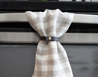 May include: A kitchen towel with a gray and white checkered pattern is held in place by a dark brown leather strap with a silver button. The towel is draped over a stainless steel oven handle.