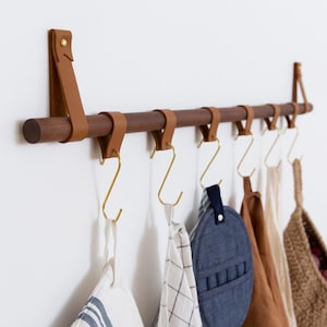 May include: A wooden wall-mounted coat rack with leather straps and gold hooks. The rack has five hooks and is holding a variety of items, including a brown tote bag, a blue hat, a white and gray striped towel, and a woven basket.