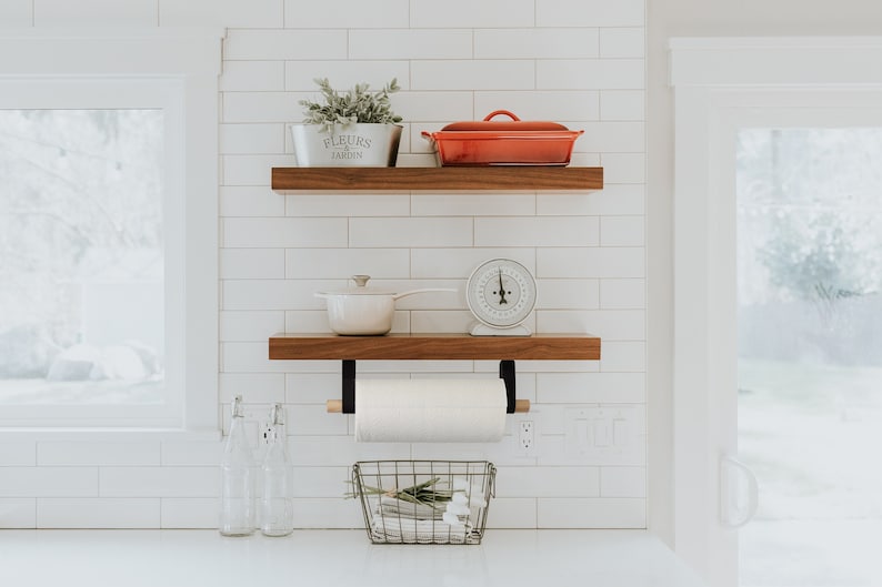 May include: Two wooden shelves with black metal brackets mounted on a white tiled wall. The top shelf has a white planter with green plants and the text 'FLEURS DU JARDIN' on it. A red ceramic baking dish is on the shelf to the right of the planter. The bottom shelf has a white pot with a handle, a white kitchen scale, and a white paper towel holder with a black metal frame. A wire basket with white flowers is on the counter below the shelves.