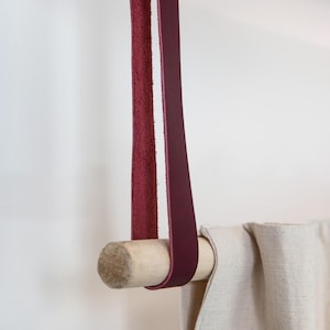 May include: A burgundy leather curtain tieback with a natural wood dowel. The leather strap is looped over the dowel and attached to the ceiling. The curtain is a light beige color.