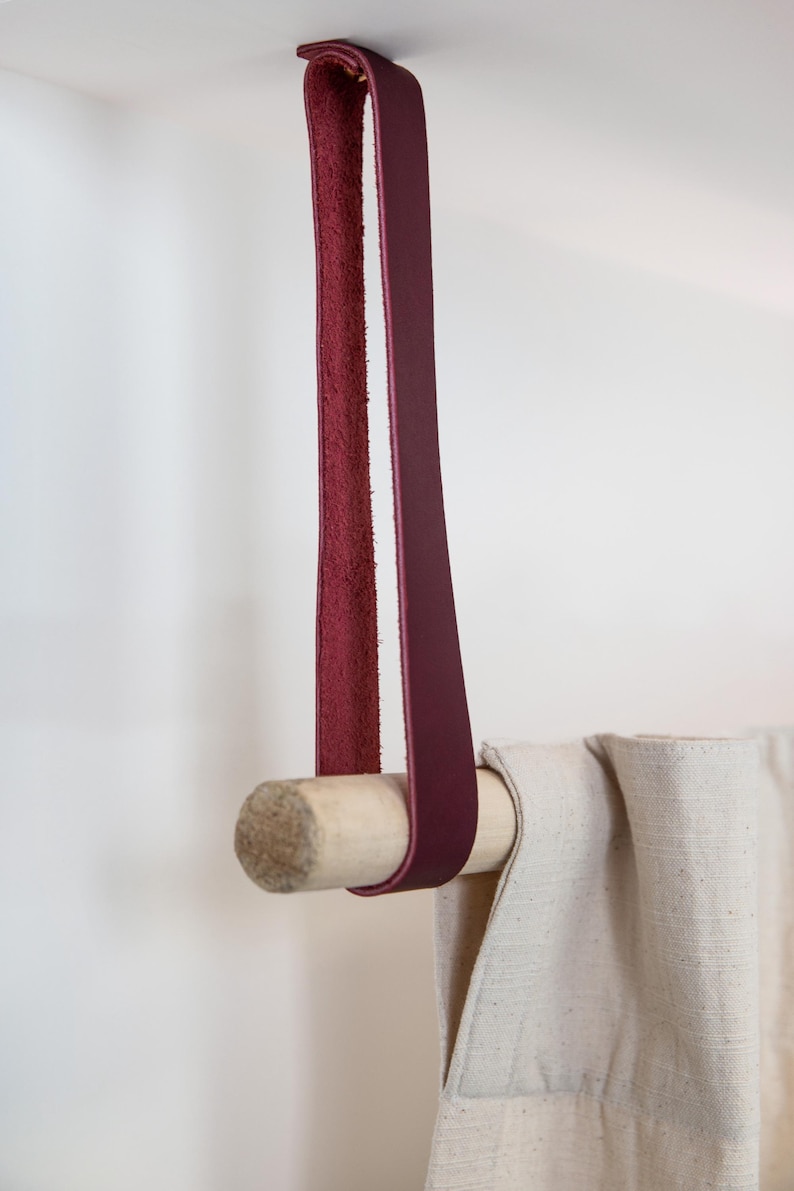 May include: A burgundy leather curtain tieback with a natural wood dowel. The leather strap is looped over the dowel and attached to the ceiling. The curtain is a light beige color.
