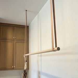 Puede incluir: Un perchero de ropa de madera clara, suspendido del techo por correas de cuero color beige. El diseño es minimalista. El fondo muestra armarios de madera y una pared blanca.