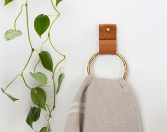May include: A gold metal towel ring with a brown leather strap attached to a white wall. A beige towel is hanging on the ring.