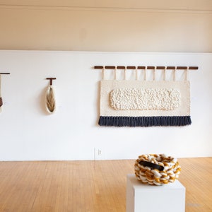 May include: A gallery wall display of woven wall hangings and baskets. The wall hangings are made of various colors and textures, including red, white, and brown. The baskets are made of natural fibers and have a variety of shapes and sizes. The text "NEST" is displayed on the wall.
