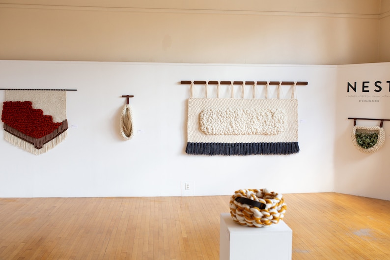 May include: A gallery wall display of woven wall hangings and baskets. The wall hangings are made of various colors and textures, including red, white, and brown. The baskets are made of natural fibers and have a variety of shapes and sizes. The text "NEST" is displayed on the wall.