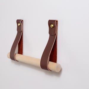 May include: A brown leather and wood toilet paper holder fixed to a white wall. The holder has two brown leather straps with gold-coloured rivets, supporting a natural wood dowel. The inside of the leather straps is reddish-brown.