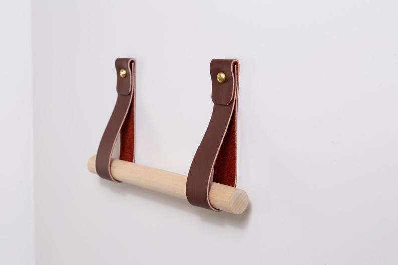 May include: A brown leather and wood toilet paper holder fixed to a white wall. The holder has two brown leather straps with gold-coloured rivets, supporting a natural wood dowel. The inside of the leather straps is reddish-brown.