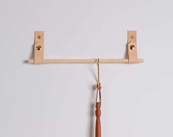 May include: A wall-mounted towel rail with a natural wood dowel and light tan leather straps secured with brass fixings. A brown leather and wood object hangs from the centre.
